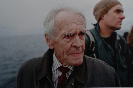 Ein älterer Mann blickt seitlich an der Kamera vorbei. Im Hintergrund ist das Meer zu sehen und ein jüngerer Mann mit einer Wollmütze. Sie befinden sich mutmaßlich auf einem Boot.