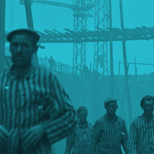 KZ-Häftlinge sind in der Bildmitte. Sie blicken direkt in die Kamera. Im Hintergrund ist die Baustelle mit Stahlträgern und Kränen sichtbar. Ein Aufseher steht am Rand des Bildes. Er blickt auf die Häftlinge. 