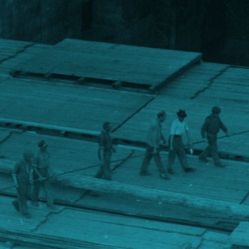 Die Aufnahme zeigt das Dach der Baustelle von oben. Die Fläche ist noch nicht fertiggestellt. Der Boden besteht auch großen Holzplatten. 18 Arbeiter in unterschiedlicher Kleidung tragen gemeinsam ein sehr lange Eisenstange auf dieser Fläche. 