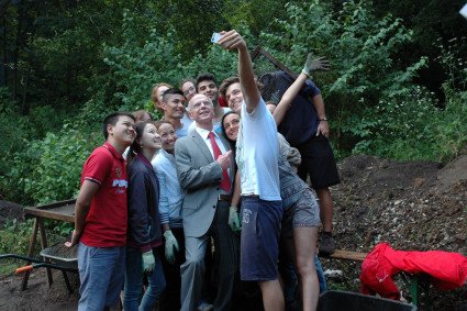 Eine Gruppe Jugendlicher hält ein Handy hoch, um ein Selfie zu erstellen mit dem Bürgerschaftspräsidenten. Die Stimmung ist ausgelassen und fröhlich. Christian Weber steht umringt von jungen Menschen unterschiedlicher Nationalitäten. Bis auf Weber tragen alle Arbeitskleidung. 