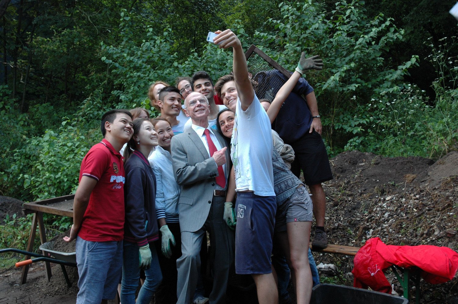Eine Gruppe Jugendlicher hält ein Handy hoch, um ein Selfie zu erstellen mit dem Bürgerschaftspräsidenten. Die Stimmung ist ausgelassen und fröhlich. Christian Weber steht umringt von jungen Menschen unterschiedlicher Nationalitäten. Bis auf Weber tragen alle Arbeitskleidung.  