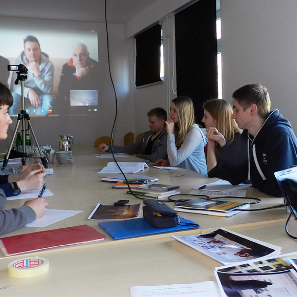 Mehrere Jugendliche sitzen gemeinsam an einem großen Konferenztisch in einem hellen Raum. Auf dem Tisch liegen Laptops, Notizblöcke, Stifte und Ausdrucke mit Fotos. An der Wand im Hintergrund ist eine Videokonferenz per Beamer zu sehen: Drei Personen sind auf einer Leinwand zu erkennen und nehmen offenbar aus der Ferne am Gespräch teil. Eine Kamera steht auf einem Stativ in der Mitte des Raums. Die Jugendlichen wirken konzentriert und arbeiten gemeinsam an einem Projekt.