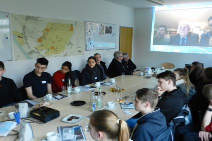 Mehrere Jugendliche sitzen gemeinsam an einem großen Konferenztisch in einem hellen Raum. Auf dem Tisch liegen Laptops, Notizblöcke, Stifte und Ausdrucke mit Fotos. An der Wand im Hintergrund ist eine Videokonferenz per Beamer zu sehen: Drei Personen sind auf einer Leinwand zu erkennen und nehmen offenbar aus der Ferne am Gespräch teil. Eine Kamera steht auf einem Stativ in der Mitte des Raums. Die Jugendlichen wirken konzentriert und arbeiten gemeinsam an einem Projekt.