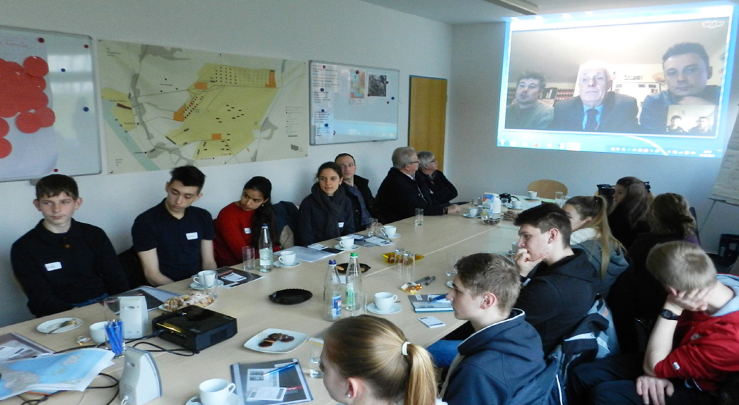 Mehrere Jugendliche sitzen gemeinsam an einem großen Konferenztisch in einem hellen Raum. Auf dem Tisch liegen Laptops, Notizblöcke, Stifte und Ausdrucke mit Fotos. An der Wand im Hintergrund ist eine Videokonferenz per Beamer zu sehen: Drei Personen sind auf einer Leinwand zu erkennen und nehmen offenbar aus der Ferne am Gespräch teil. Eine Kamera steht auf einem Stativ in der Mitte des Raums. Die Jugendlichen wirken konzentriert und arbeiten gemeinsam an einem Projekt. 