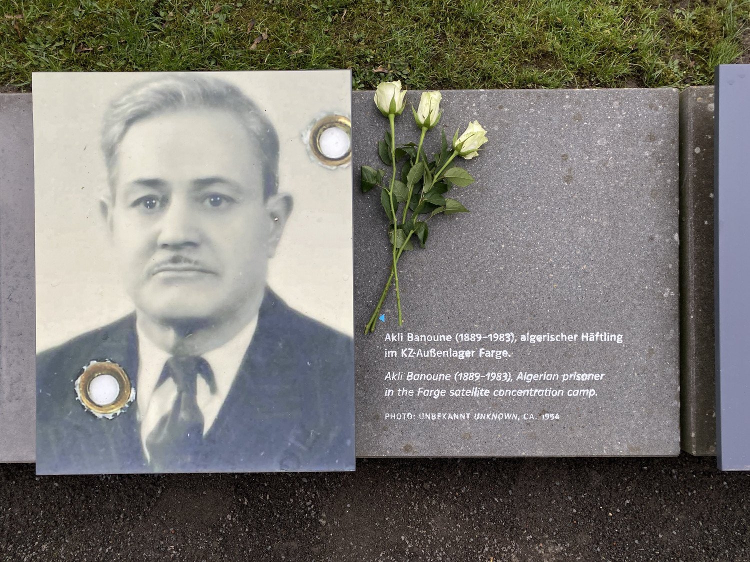 Gedenkstein am Wegesrand mit einem Foto von Akli Banoune (1889 - 1983) und einem kurzen Text: Algerischer Häftling im KZ-Außenlager Farge. Auf der Steintafel liegen drei weiße Rosen.  