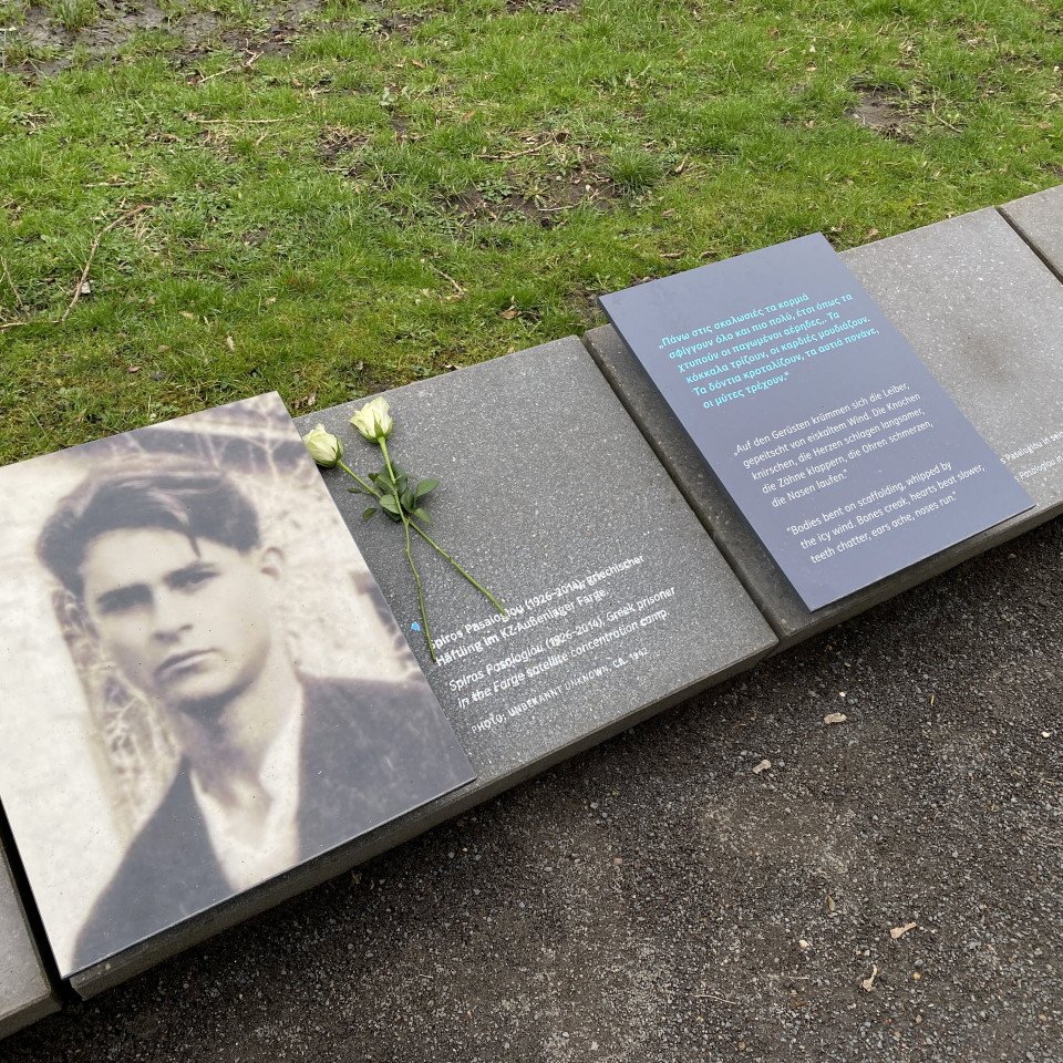 Gedenkstein am Wegesrand mit einem Foto von Spiros Pasaloglou und einem kurzen Text mit seinen Lebensdaten sowie einem Zitat in deutsch und englisch. Auf dem Gedenkstein liegen zwei weiße Rosen.
