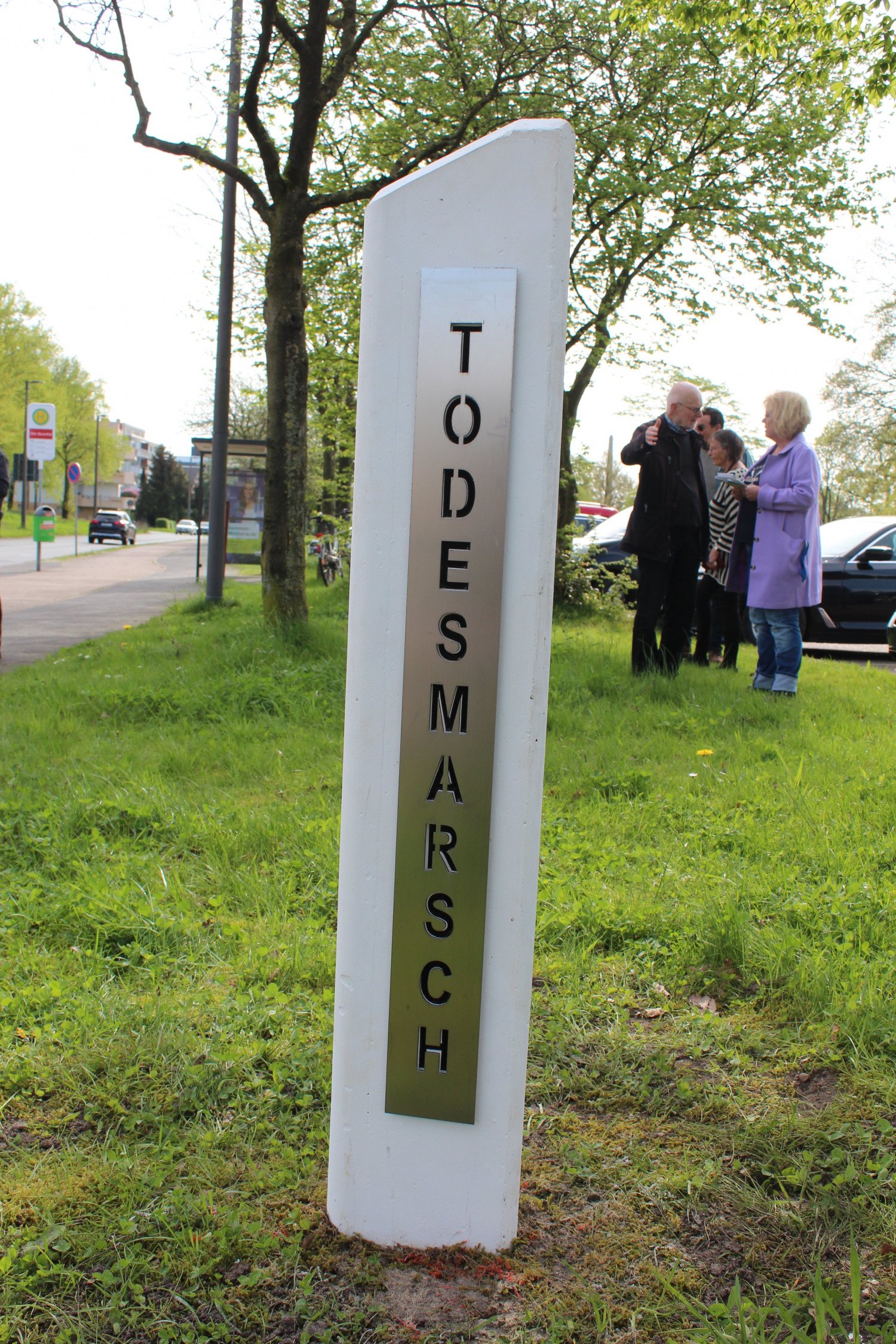 Weiße Stele, die mit dem Wort Todesmarsch beschriftet ist. Die Stele befindet sich in einer Grünanlage. Im Bildhintergrund stehen Menschen, die miteinander sprechen.  