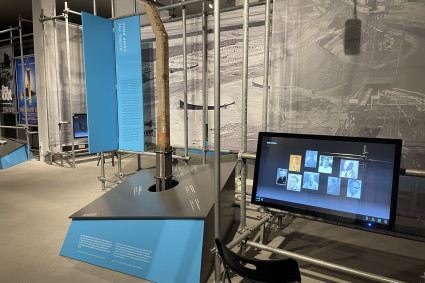 Das Foto zeigt einen modernen Museumsraum mit einer Ausstellung. Im Mittelpunkt steht eine Installation aus Metallrohren und blauen Infotafeln. In der Mitte ist ein längliches, rostfarbenes Objekt – vermutlich Zaunpfosten. Daneben befindet sich eine schwarze Informationsfläche mit weißer Schrift, die in den Ausstellungstisch integriert ist. Rechts im Bild steht ein großer Bildschirm, auf dem Porträtfotos von mehreren Personen zu sehen sind. Vor dem Bildschirm steht ein schwarzer Stuhl. Im Hintergrund erkennt man eine großformatige Schwarz-Weiß-Fotografie, die eine Baustelle oder ein Industriegelände zeigt. Der Raum ist hell ausgeleuchtet und wirkt technisch-modern gestaltet. Ein blauer Textabschnitt auf der linken Seite trägt den englischen Titel „Along the Camp Road“ (Deutsch: „Entlang der Lagerstraße“).