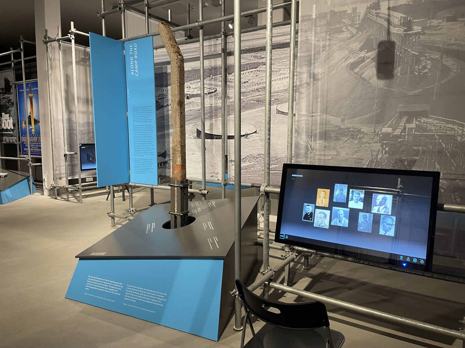 Das Foto zeigt einen modernen Museumsraum mit einer Ausstellung. Im Mittelpunkt steht eine Installation aus Metallrohren und blauen Infotafeln. In der Mitte ist ein längliches, rostfarbenes Objekt – vermutlich Zaunpfosten. Daneben befindet sich eine schwarze Informationsfläche mit weißer Schrift, die in den Ausstellungstisch integriert ist. Rechts im Bild steht ein großer Bildschirm, auf dem Porträtfotos von mehreren Personen zu sehen sind. Vor dem Bildschirm steht ein schwarzer Stuhl. Im Hintergrund erkennt man eine großformatige Schwarz-Weiß-Fotografie, die eine Baustelle oder ein Industriegelände zeigt. Der Raum ist hell ausgeleuchtet und wirkt technisch-modern gestaltet. Ein blauer Textabschnitt auf der linken Seite trägt den englischen Titel „Along the Camp Road“ (Deutsch: „Entlang der Lagerstraße“). 