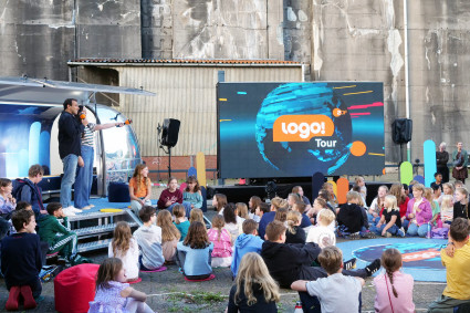 Im Vordergrund sitzen zahlreiche Kinder in lockeren Reihen auf dem Boden und auf Kissen. Sie schauen nach vorne zu einer kleinen Bühne. Auf dieser Bühne stehen zwei Personen mit Mikrofonen und sprechen ins Publikum. Links hinter ihnen steht ein silbernes, rundes Fahrzeug mit offener Seite – das „logo!“-Mobil. Rechts daneben befindet sich eine große LED-Leinwand. Darauf ist das blaue „logo!“-Weltkugel mit dem Schriftzug „logo! Tour ZDF“ zu sehen. Im Hintergrund erkennt man eine hohe graue Betonwand und ein flaches Gebäude mit Wellblechdach. Einige Erwachsene stehen rechts am Rand und beobachten die Szene. Die Stimmung wirkt lebendig und fröhlich, passend zu einer Kinderveranstaltung im Freien.