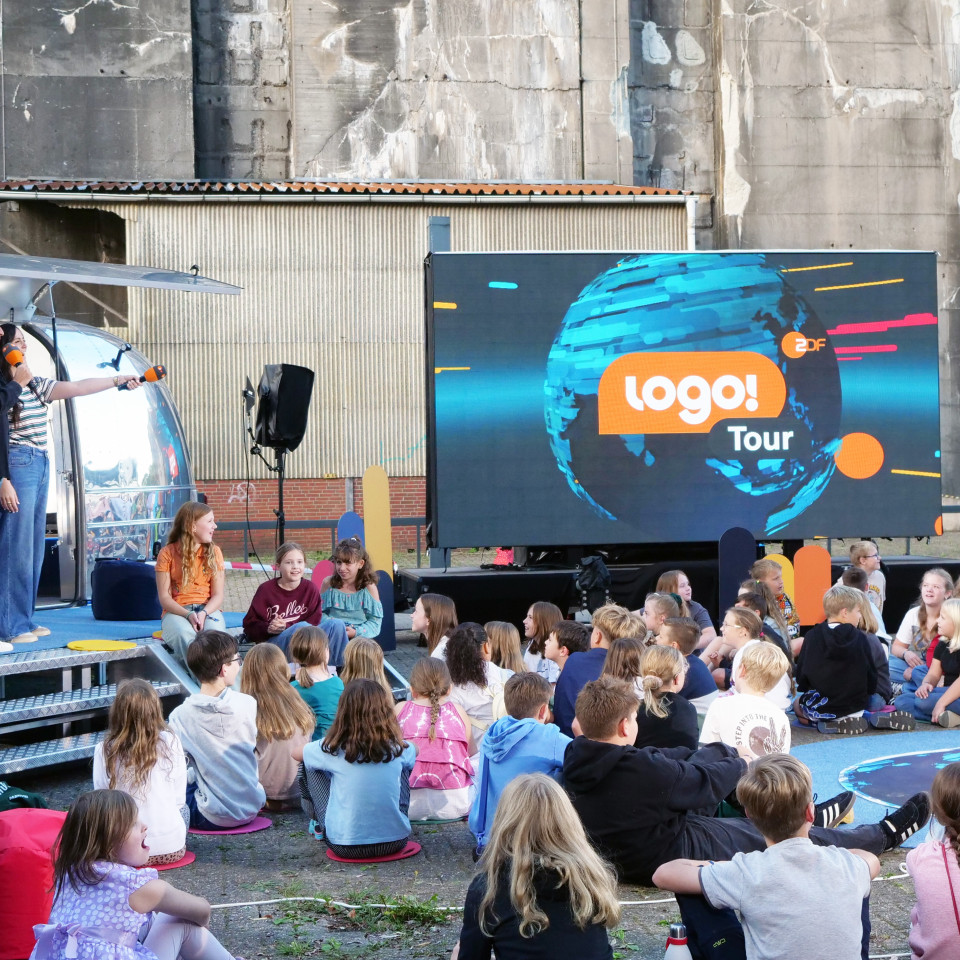Im Vordergrund sitzen zahlreiche Kinder in lockeren Reihen auf dem Boden und auf Kissen. Sie schauen nach vorne zu einer kleinen Bühne. Auf dieser Bühne stehen zwei Personen mit Mikrofonen und sprechen ins Publikum. Links hinter ihnen steht ein silbernes, rundes Fahrzeug mit offener Seite – das „logo!“-Mobil. Rechts daneben befindet sich eine große LED-Leinwand. Darauf ist das blaue „logo!“-Weltkugel mit dem Schriftzug „logo! Tour ZDF“ zu sehen. Im Hintergrund erkennt man eine hohe graue Betonwand und ein flaches Gebäude mit Wellblechdach. Einige Erwachsene stehen rechts am Rand und beobachten die Szene. Die Stimmung wirkt lebendig und fröhlich, passend zu einer Kinderveranstaltung im Freien.