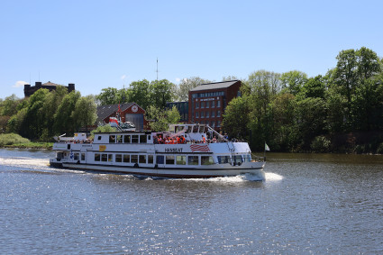 Ein Ausflugsschiff fährt auf einem ruhigen Fluss, umgeben von Bäumen und Gebäuden, bei sonnigem Wetter.