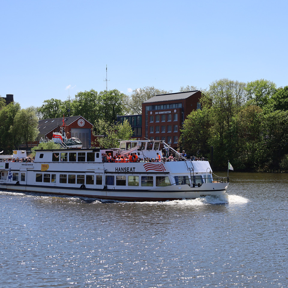 Ein Ausflugsschiff fährt auf einem ruhigen Fluss, umgeben von Bäumen und Gebäuden, bei sonnigem Wetter.