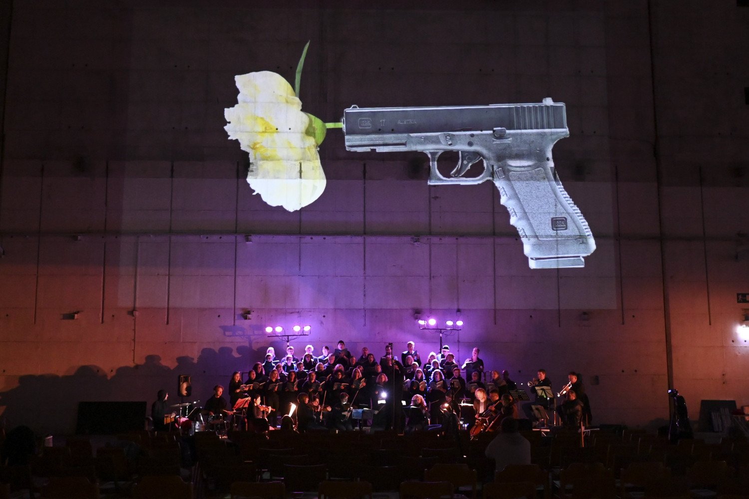 Es ist eine Pistole auf die Bunkerwand projiziert. Aus ihr entspringt eine Blumen-Blüte. Konzert-Musiker:innen sitzen darunter.  