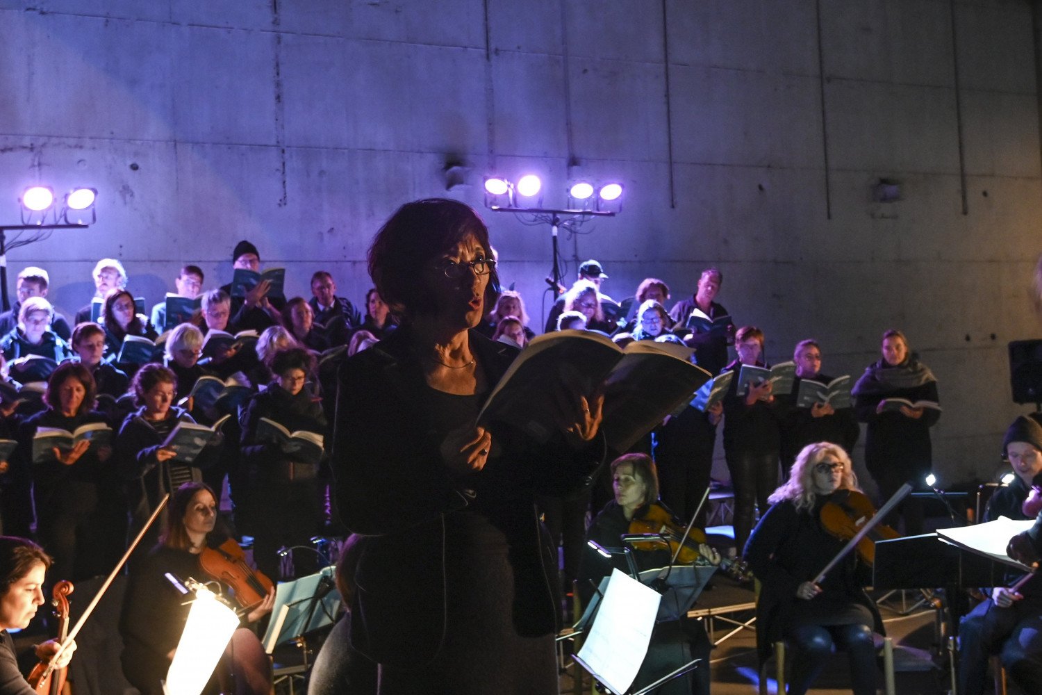 Eine Sängerin mit Textbuch inmitten von Konzert-Musiker:innen.   