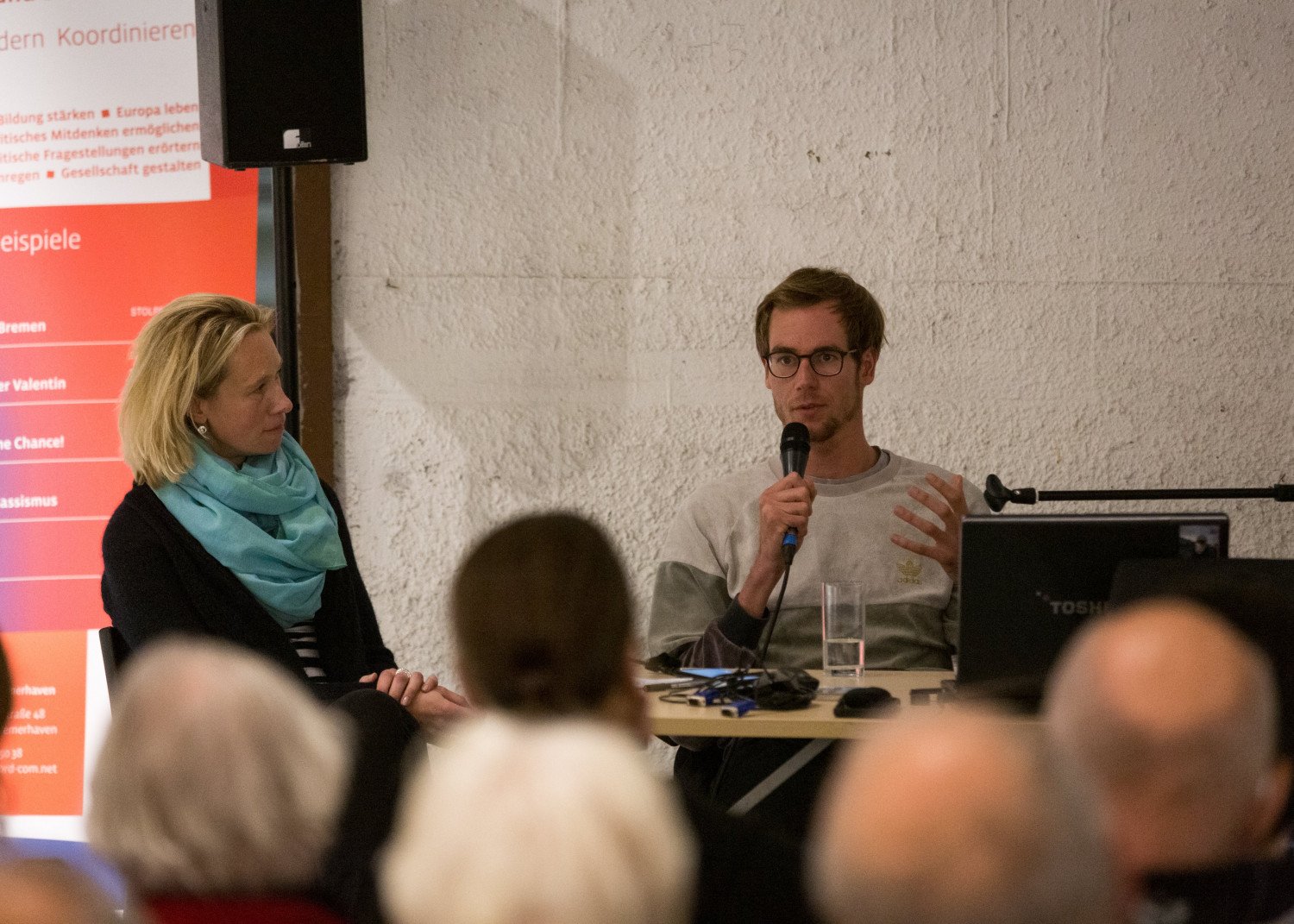 Der Autor und die Moderatorin im Gespräch. Der Autor hält ein Mikrophon in der Hand. Vor ihnen sitzt Publikum.  