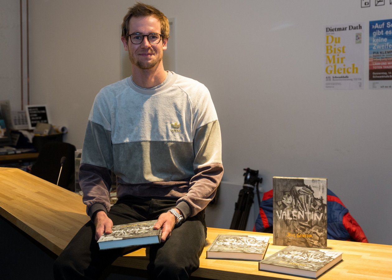 Der Autor sitzt auf einem Infotresen und hält sein Buch in der Hand. Neben ihm liegen weitere Exemplare von "Valentin".