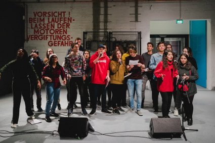 20 Jungendliche Singen und Performen im Infozentrum des Denkort. Sie sind bunt gekleidet. Halten Mikros und Textblätter in der Hand. 