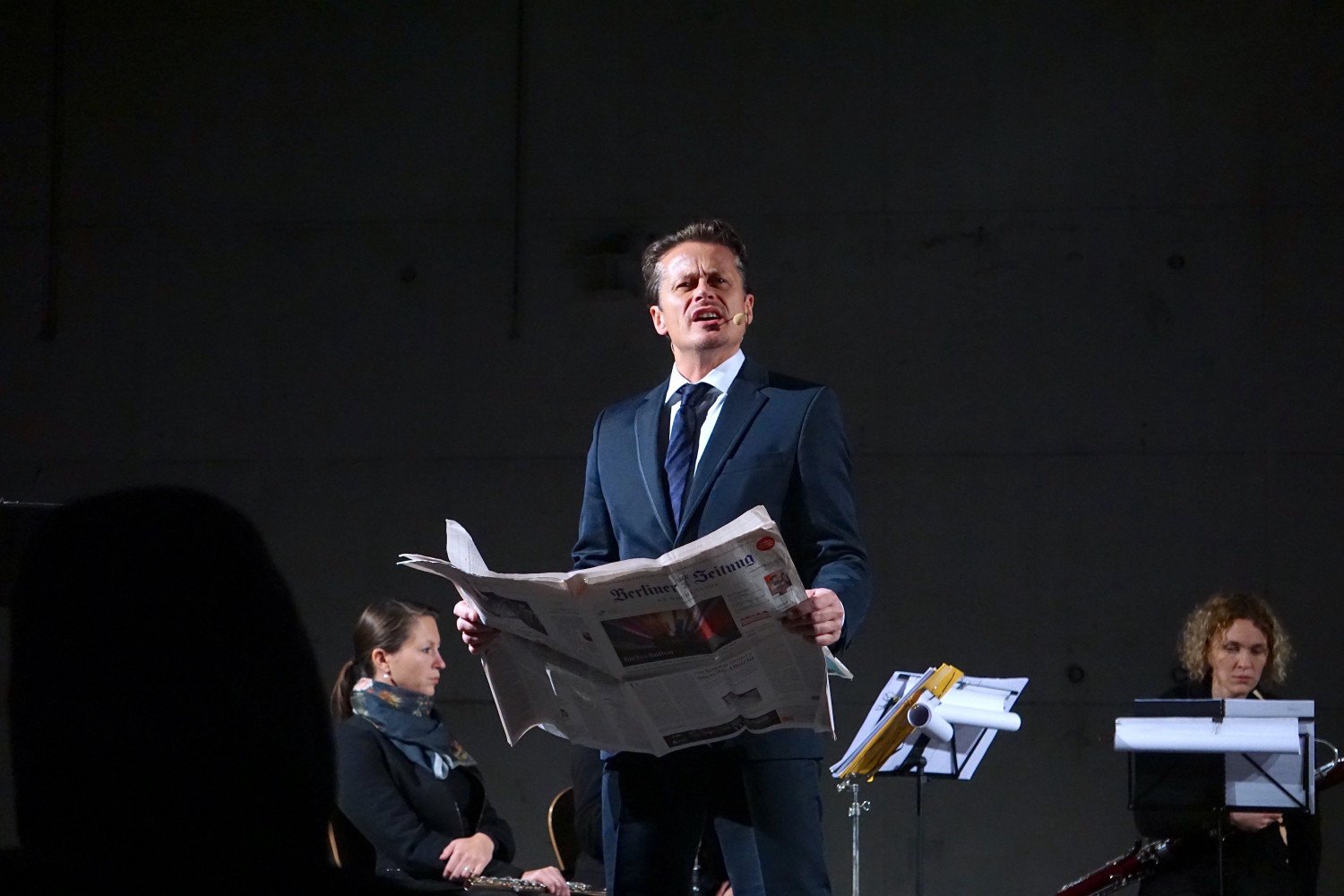 Ein Schauspieler mit einer Zeitung in der Hand. Die Zeitung ist geöffnet. Er trägt daraus vor.  