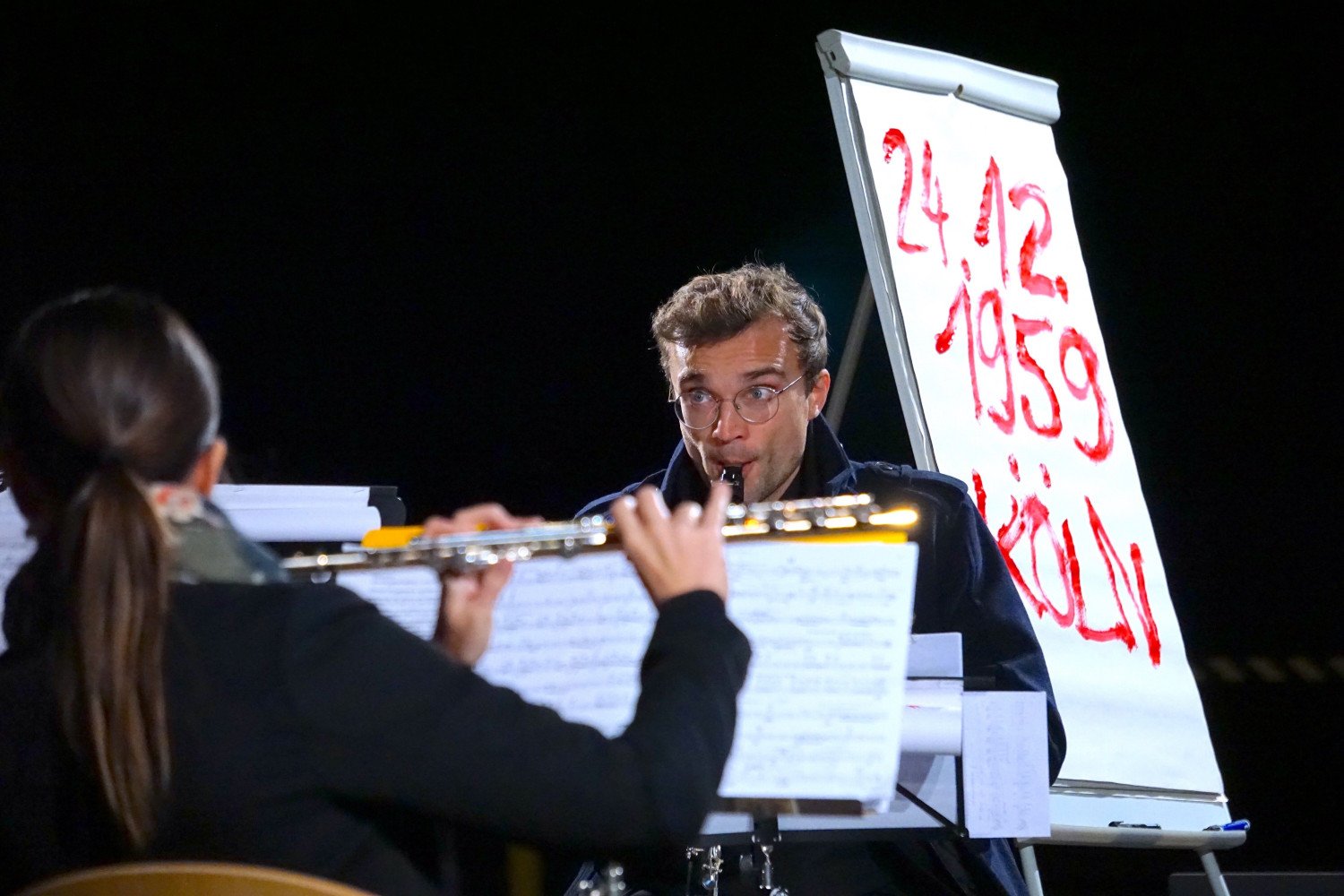 Zwei Musiker:innen spielend. Dahinter das Plakat mit roter Schrift. 