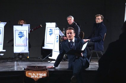 Drei Musiker:innen spielend. Vor ihnen jeweils ein Plakat mit den Worten Münchener Oktoberfest und einer weiss-blauen Abbildung. Mit dem Gesicht in die Kamera gerichtet blickt der Schauspieler das Publikum an. Er kniet. Vor ihm ist eine Aktentasche mit dem Datum 11.4.1968 aufgetragen.