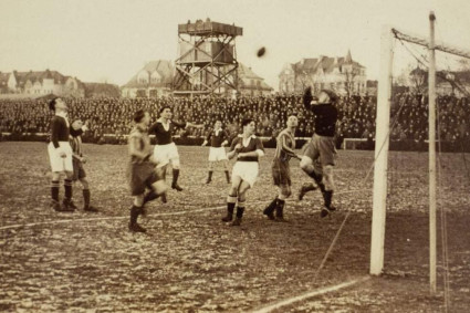 Schwarzweißes historisches Foto eines Fußballspiels in einem großen Stadion. Im Vordergrund stehen mehrere Spieler dicht vor dem Tor. Ein Torwart springt nach oben und greift mit ausgestreckten Armen nach dem Ball, während andere Spieler um ihn herum stehen oder zum Ball schauen. Das Tor mit Netz ist rechts im Bild zu sehen.<br />
Im Hintergrund befindet sich eine sehr große Menschenmenge auf den Tribünen. Hinter den Zuschauern stehen Gebäude sowie ein erhöhter Turm oder eine Plattform.