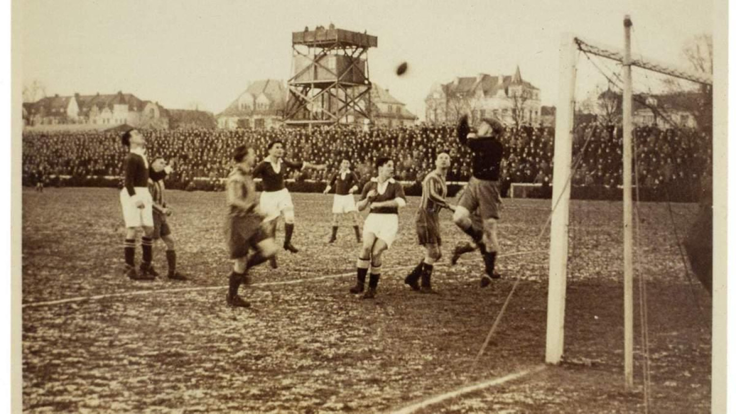 Schwarzweißes historisches Foto eines Fußballspiels in einem großen Stadion. Im Vordergrund stehen mehrere Spieler dicht vor dem Tor. Ein Torwart springt nach oben und greift mit ausgestreckten Armen nach dem Ball, während andere Spieler um ihn herum stehen oder zum Ball schauen. Das Tor mit Netz ist rechts im Bild zu sehen.<br />
Im Hintergrund befindet sich eine sehr große Menschenmenge auf den Tribünen. Hinter den Zuschauern stehen Gebäude sowie ein erhöhter Turm oder eine Plattform. 
