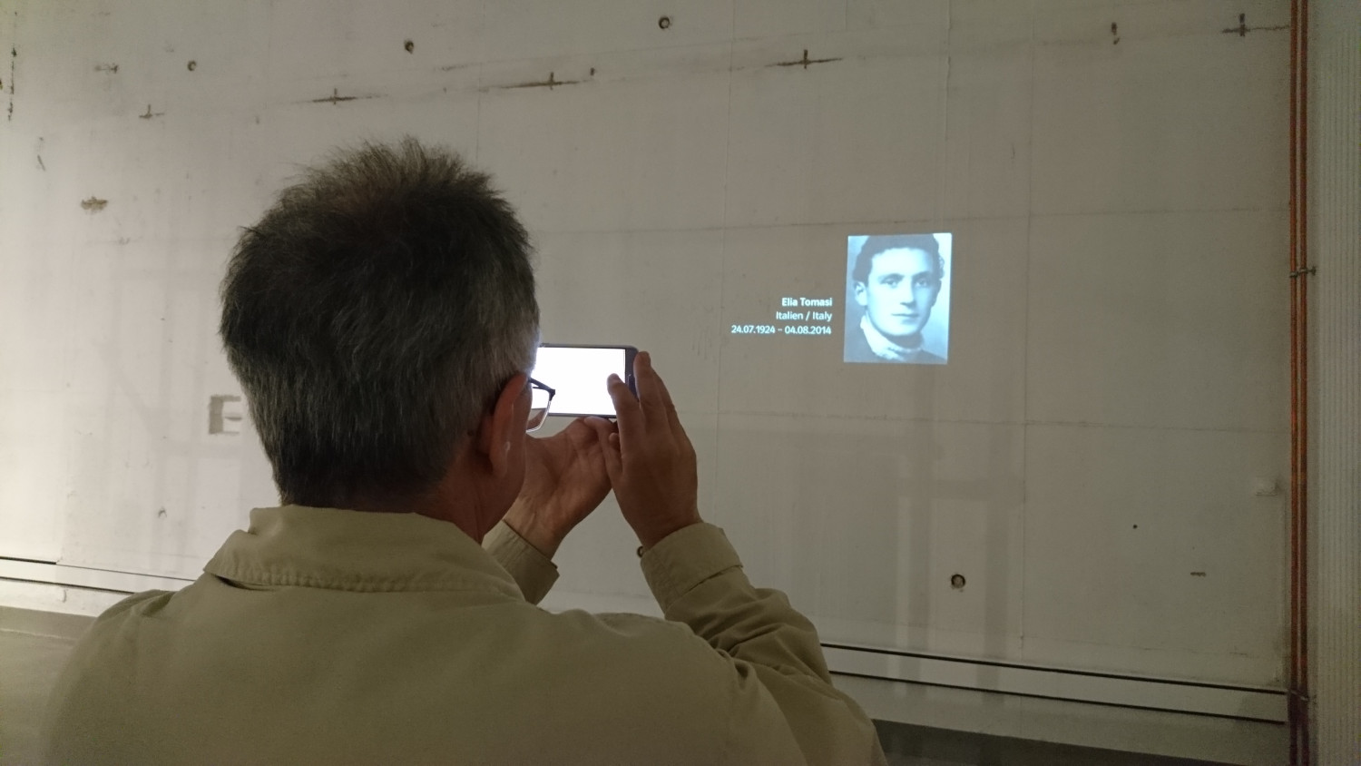 Eine Mann steht vor einer Wand und fotografiert ein projiziertes Portrait mit Informationen zu einer abgebildeten Person. 