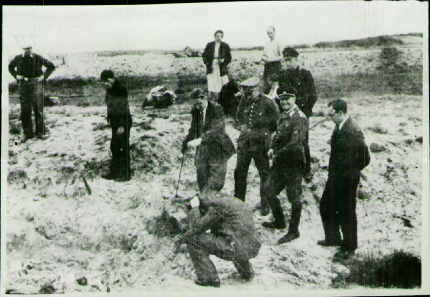 Abgebildet wird ein schwarz-weißes Bild in sehr schlechter Qualität. Es sind 9 Männer und eine Frau auf dem Bild zu sehen. Im Vordergrund ist ein Mann zu sehen, der in der Hocke sitzt und den Fundort der Gräber fotografiert. Er ist ein Beamter der Stader Kripo. Neben ihm steht ein Mann, der in die Kamera lächelt. Die anderen Personen beobachten den Prozess.  