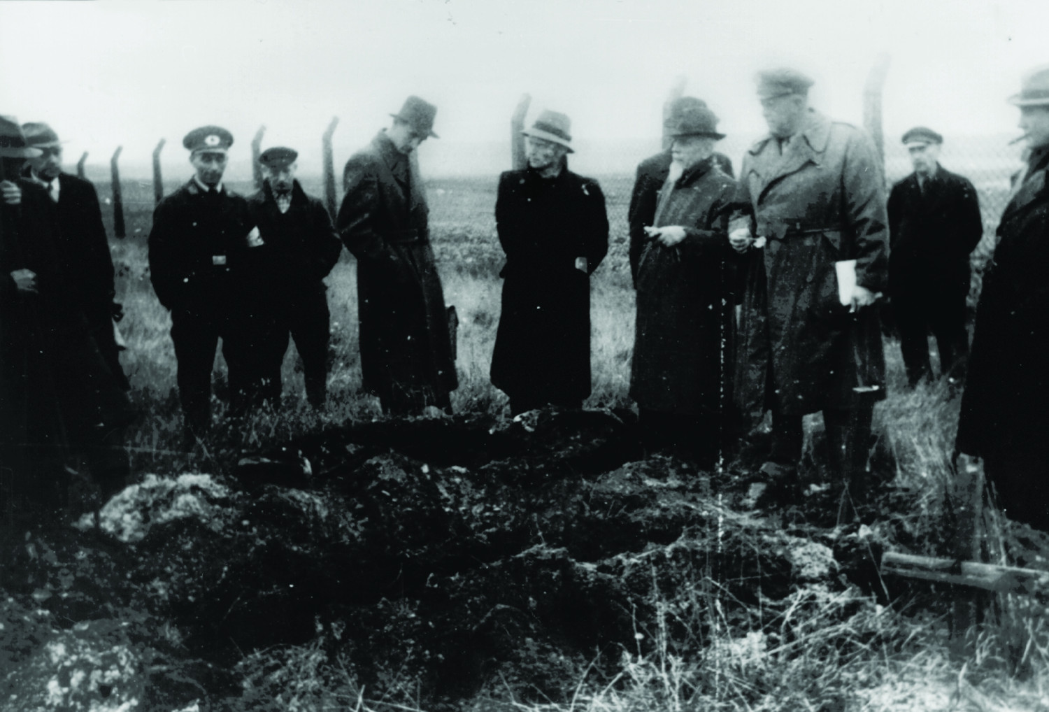 Gezeigt werden 11 Männer auf einem schwarz-weißen Bild. Sie stehen vor einem ausgehobenem Gräberfeld. Im Hintergrund sieht man einen Zaun. Das Gräberfeld ist durch ein aus Stöcken zusammengebundenem Holzkreuz im Vordergrund gekennzeichnet. Die Männer tragen mutmaßlich alle schwarze Kleidung und Mäntel sowie Kopfbedeckung.  