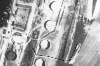 Abgebildet ist eine Luftaufnahme in schwarz-weiß. Sie zeigt anhand einer nachträglich eingefügten schwarzen Linie den Sandweg, der zum Gräberfeld der Rekumer Feldmark führte. Man sieht auf der Luftaufnahme ansonsten vor allem Felder und große Kreise, die vermutlich Öltanks oder Silos darstellen. 