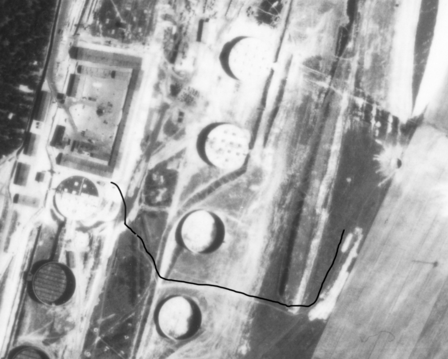 Abgebildet ist eine Luftaufnahme in schwarz-weiß. Sie zeigt anhand einer nachträglich eingefügten schwarzen Linie den Sandweg, der zum Gräberfeld der Rekumer Feldmark führte. Man sieht auf der Luftaufnahme ansonsten vor allem Felder und große Kreise, die vermutlich Öltanks oder Silos darstellen.  