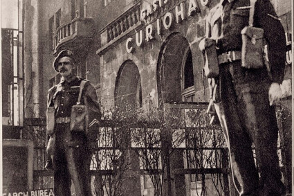 Es ist ein sepiafarbenes Bild abgebildet. Im Vordergrund stehen zwei Wachmänner, im Hintergrund ist die Gaststätte Curiohaus zu sehen. Im linken unteren Bildrand steht abgeschnitten: englisch "Search Bureau Control Commission" mit einem Pfeil nach rechts. Die beiden Männer schauen an der Kamera vorbei, der Blick ist sehr ernst. Vor dem Gebäude des Curiohauses sind kahle Sträucher zu sehen. 