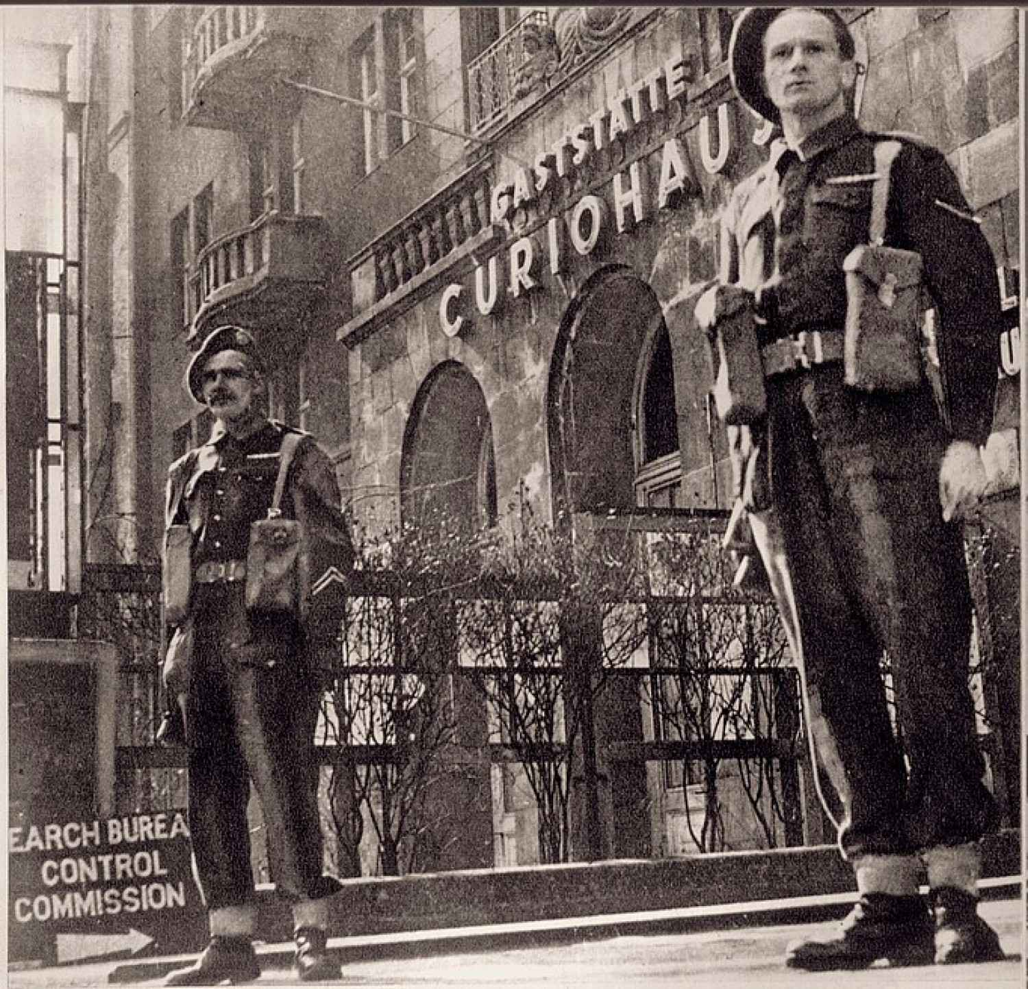 Es ist ein sepiafarbenes Bild abgebildet. Im Vordergrund stehen zwei Wachmänner, im Hintergrund ist die Gaststätte Curiohaus zu sehen. Im linken unteren Bildrand steht abgeschnitten: englisch "Search Bureau Control Commission" mit einem Pfeil nach rechts. Die beiden Männer schauen an der Kamera vorbei, der Blick ist sehr ernst. Vor dem Gebäude des Curiohauses sind kahle Sträucher zu sehen.  