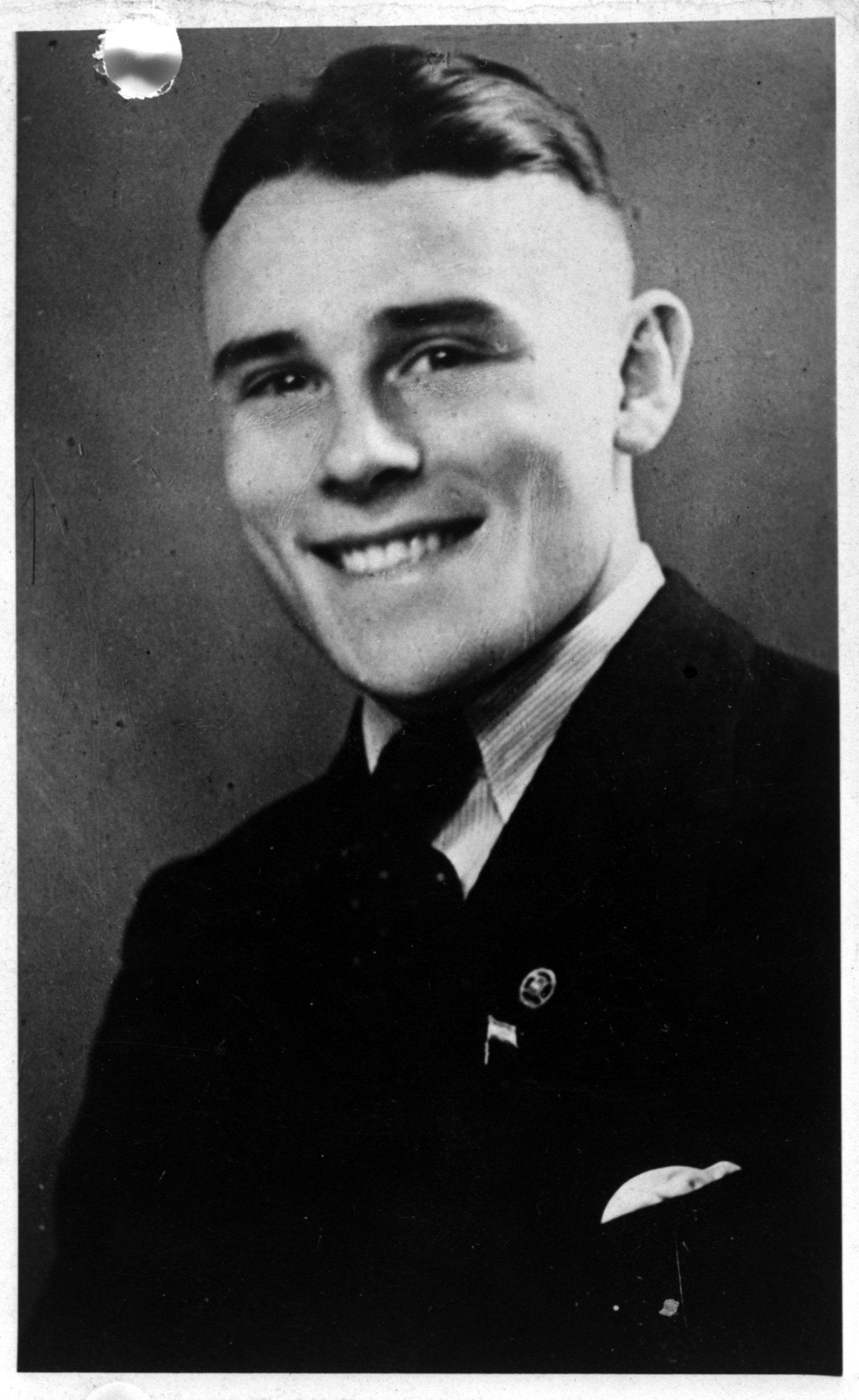 Abgebildet ist ein schwarz-weißes Portrait von einem lachenden Mann mit gegelten Haaren. Es handelt sich um Heinrich Schauwacker. Er trägt einen Anzug und eine Krawatte. Der Blick ist in die Kameralinse gerichtet und wirkt stolz.  