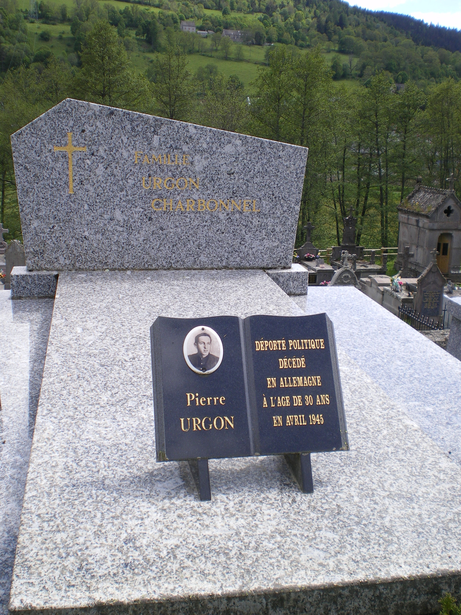 Abgebildet ist ein Foto von einem Grabstein der Familie Urgon, im Vordergrund ist die Gedenktafel mittig auf dem Grab zu erkennen, im Hintergrund des Bildes sieht man weitere Gräber sowie Bäume und eine Wiese.  