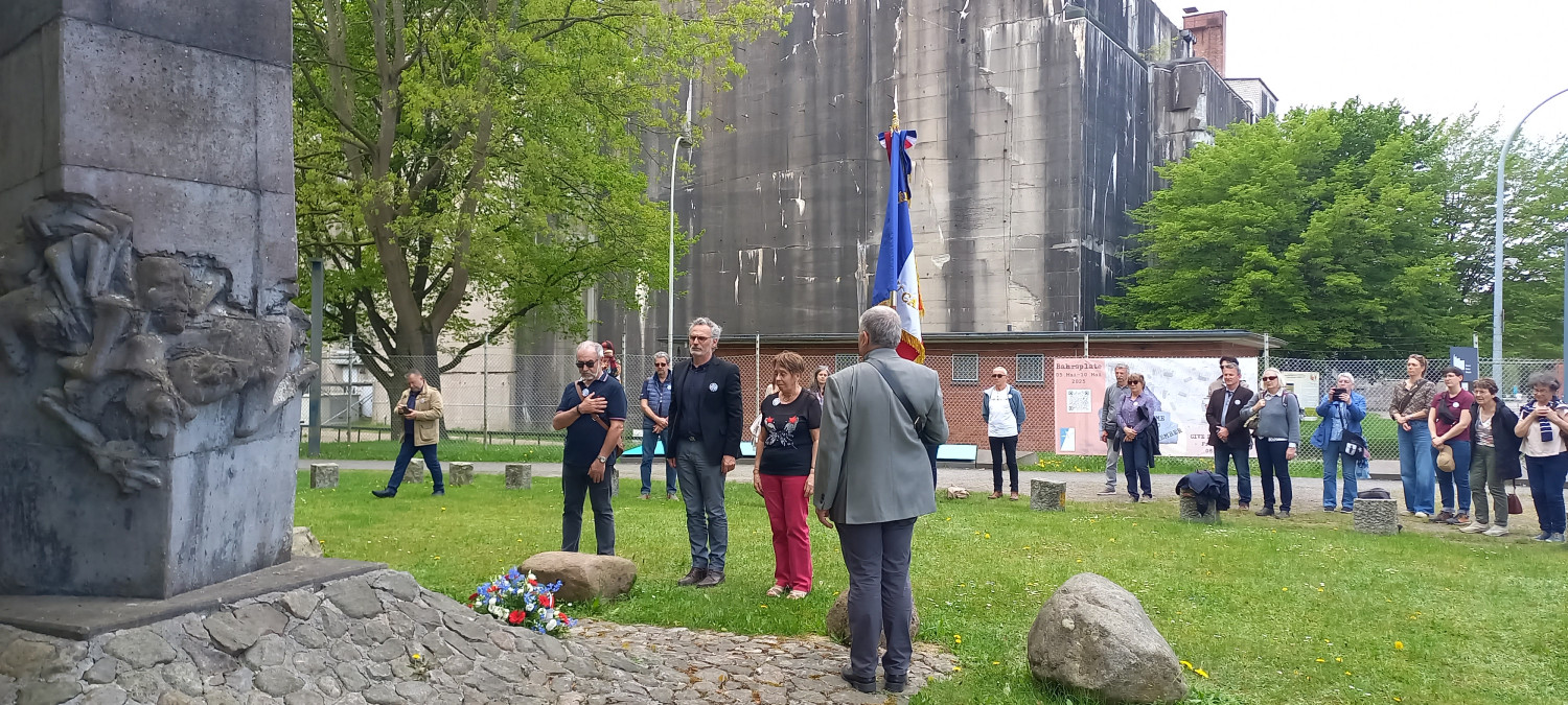 Gezeigt wird ein Foto, links im Bild ist ein Mahnmal für Zwangsarbeiter:innen zu sehen. Daneben stehen eine Frau und drei Männer, die den Blick auf einen Kranz vor ihren Füßen gerichtet haben. Der ganz links stehende Mann hat die rechte Hand auf das Herz gelegt. Ein Mann steht mit dem Rücken zur Kamera, er trägt eine französische Fahne. Dahinter stehen mehrere Personen und beobachten die mittig stehenden Menschen.  