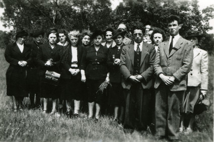 Gezeigt wird ein schwarz-weißes Bild, es zeigt 12 Frauen und 3 Männer. Die Frauen sind überwiegend in schwarz gekleidet, die Männer tragen graue Anzüge. Im Hintergrund befindet sich ein Baum, die Personen stehen auf einer Wiese.