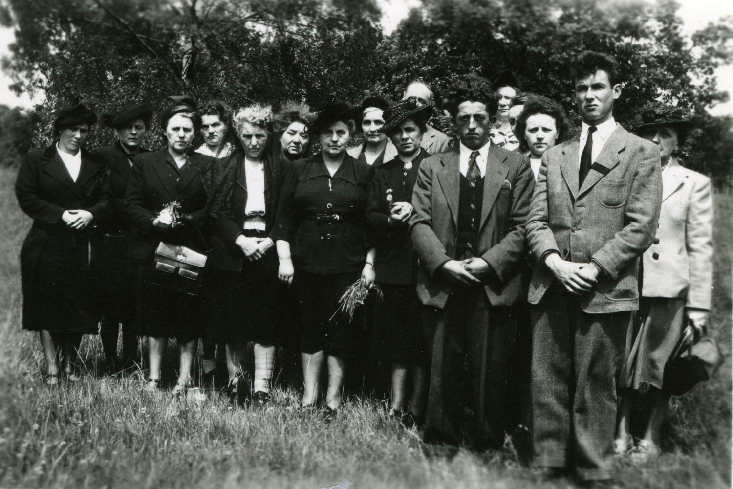 Gezeigt wird ein schwarz-weißes Bild, es zeigt 12 Frauen und 3 Männer. Die Frauen sind überwiegend in schwarz gekleidet, die Männer tragen graue Anzüge. Im Hintergrund befindet sich ein Baum, die Personen stehen auf einer Wiese. 