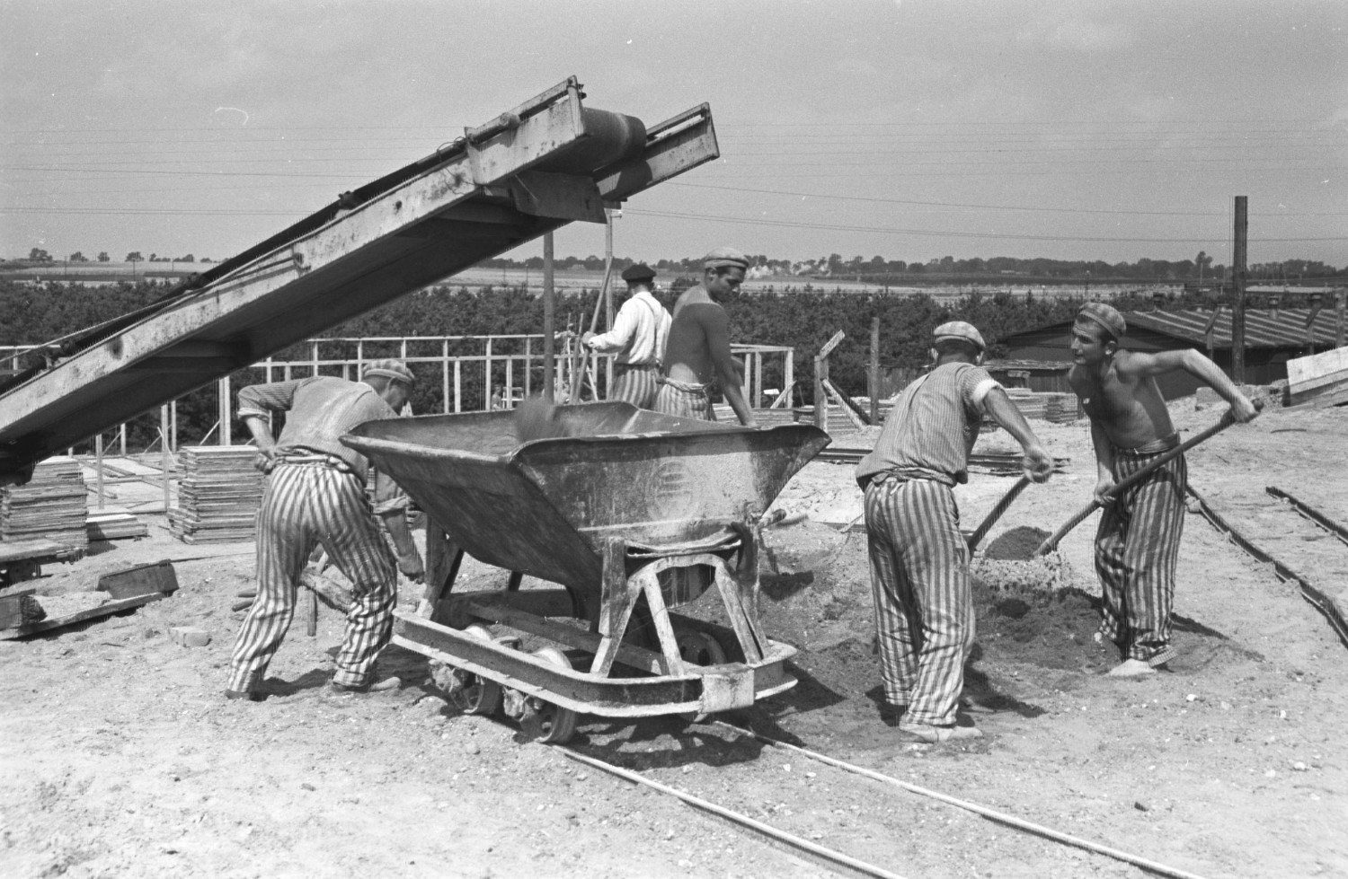 Historische Aufnahme in schwarz-weiss aus dem Jahr 1944. Fünf Männer in gestreifter Häftlingsbekleidung schippen Sand in einen auf Schienen stehenden Container. Darüber befindet sich ein Laufband. Im Hintergrund lagern weitere Baumaterialien. 