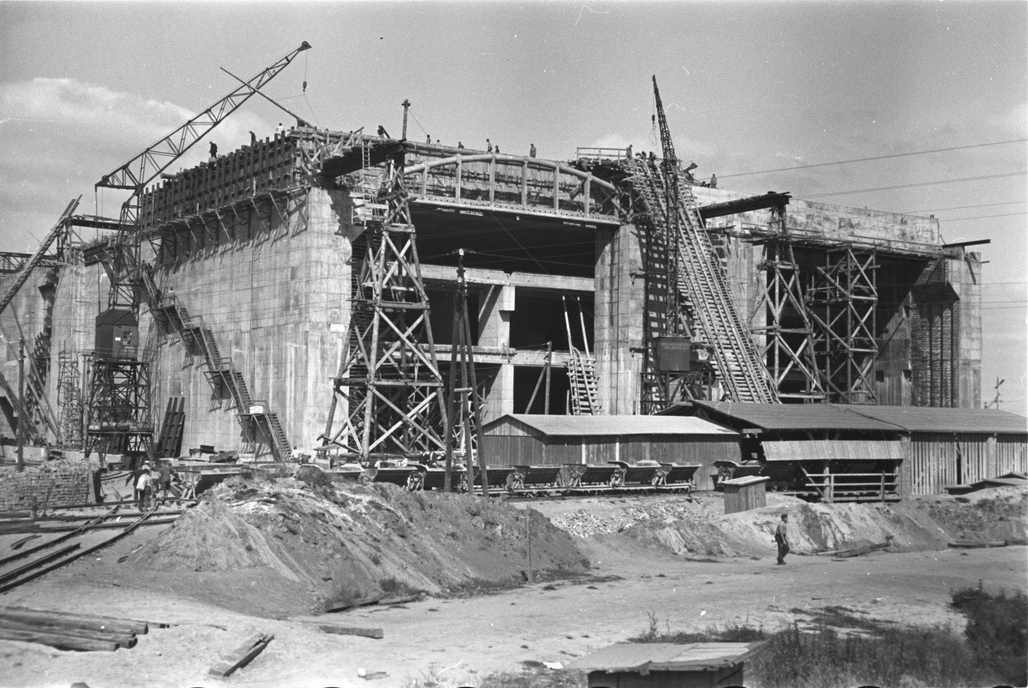 Historische Aufnahme in schwarz-weiss aus dem Jahr 1944. Die Bunkeranlage ist von Gerüsten umgeben. Auf dem Dach und den Wegen herum erkennt man Menschen. Diese wirken im Vergleich zum Gebäude winzig. Um den Bunker herum befinden sich große Sandberge, einfach Holzbaracken und Schienen mit Bahnwaggons darauf. 