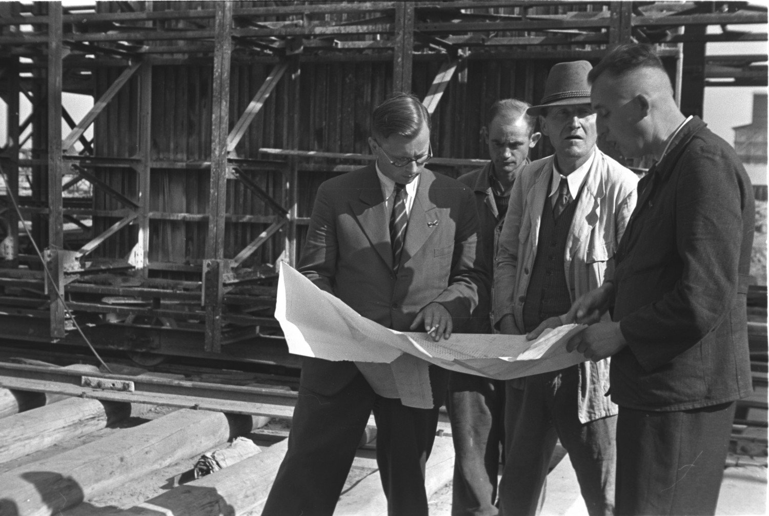 Historische Aufnahme in schwarz-weiss aus dem Jahr 1944. Vier Männer stehen auf der Baustelle und blicken auf einen Plan, den sie aufgespannt in ihren Händen halten. Sie tragen gepflegte Kleidung, zum Teil mit Krawatte. Einer von ihnen trägt einen Herrenhut. Es handelt sich um den Bauleiter Steig. Es scheint sich um eine Besprechung zu handeln. .  