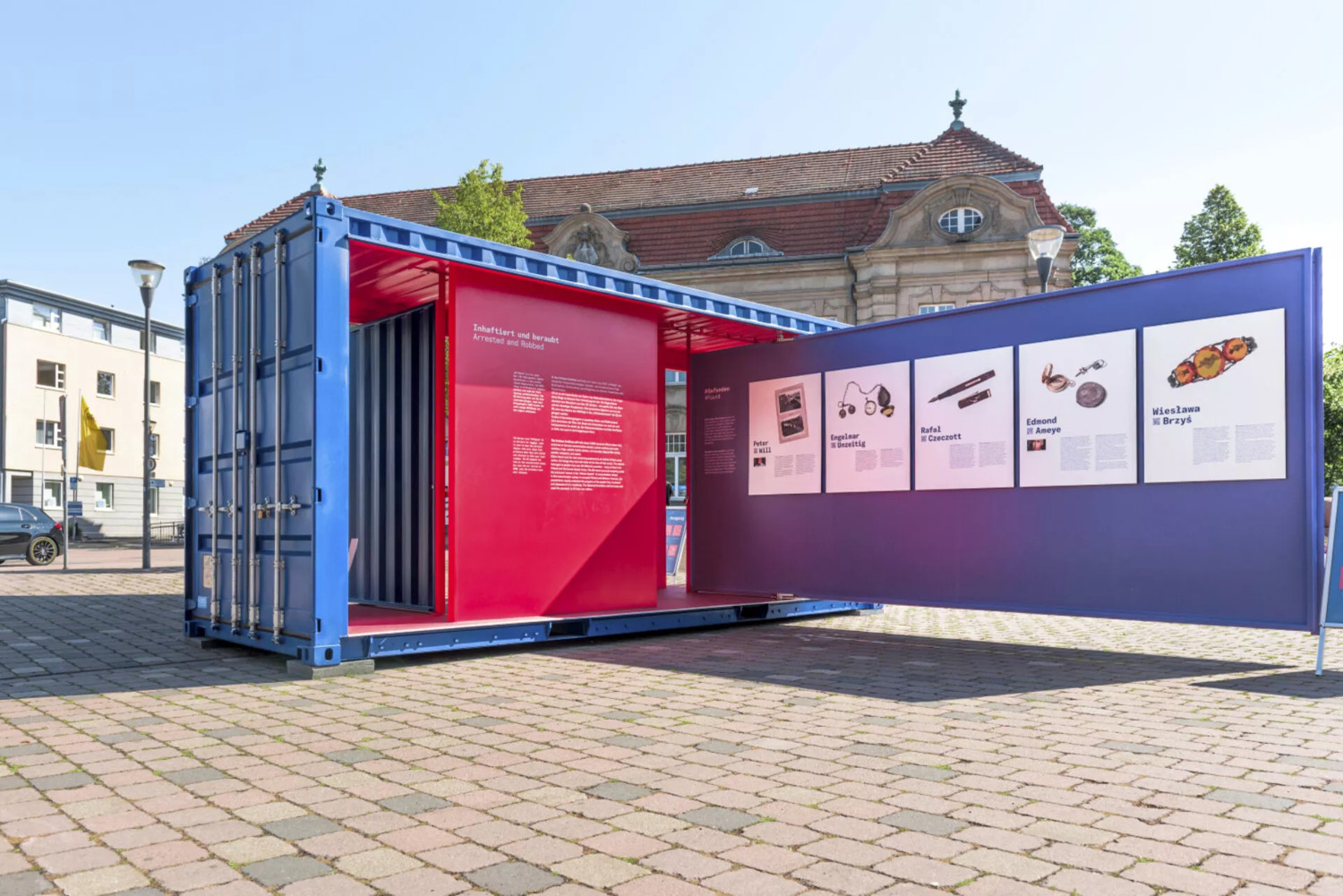 Gezeigt wird ein blauer, geöffneter Container. Er ist begehbar und mit einer Ausstellung versehen, die im Kontrast zur Farbe des Containers mit rot gestaltet wurde. Die offene Tür des Containers zeigt Fundstücke, die aus der Zeit des Nationalsozialismus stammen. Sie wurden Menschen, die verschiedene Lagersysteme des zweiten Weltkriegs durchlaufen haben, abgenommen. Im Container selbst sieht man einen Schriftzug: "Inhaftiert und beraubt".  