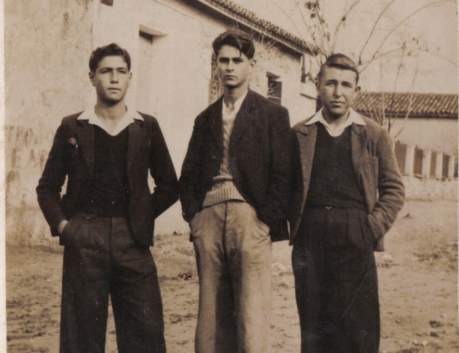 Abgebildet ist ein sepiafarbenes Bild. Es zeigt drei junge Männer, welche für das Foto posieren. Sie blicken ernst in die Kamera. Im Hintergrund sind Häuser und Bäume zu erkennen. 