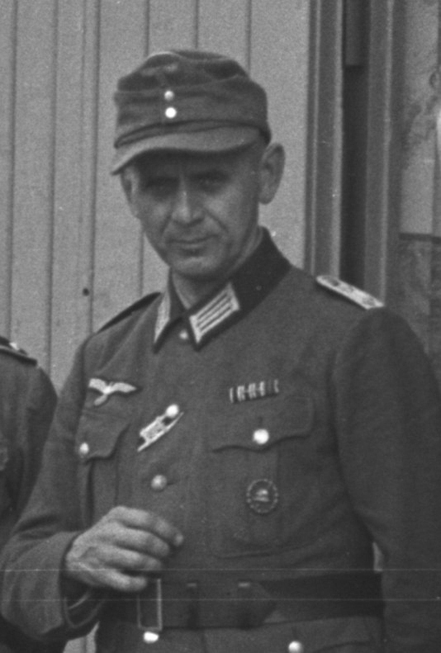 Gezeigt wird ein schwarz-weißes Bild. Zu sehen ist ein uniformierter Mann, der in die Kamera blickt. Den rechten Arm hat er etwas angewinkelt und hält ihn über seinem Gürtel am Bauch. 