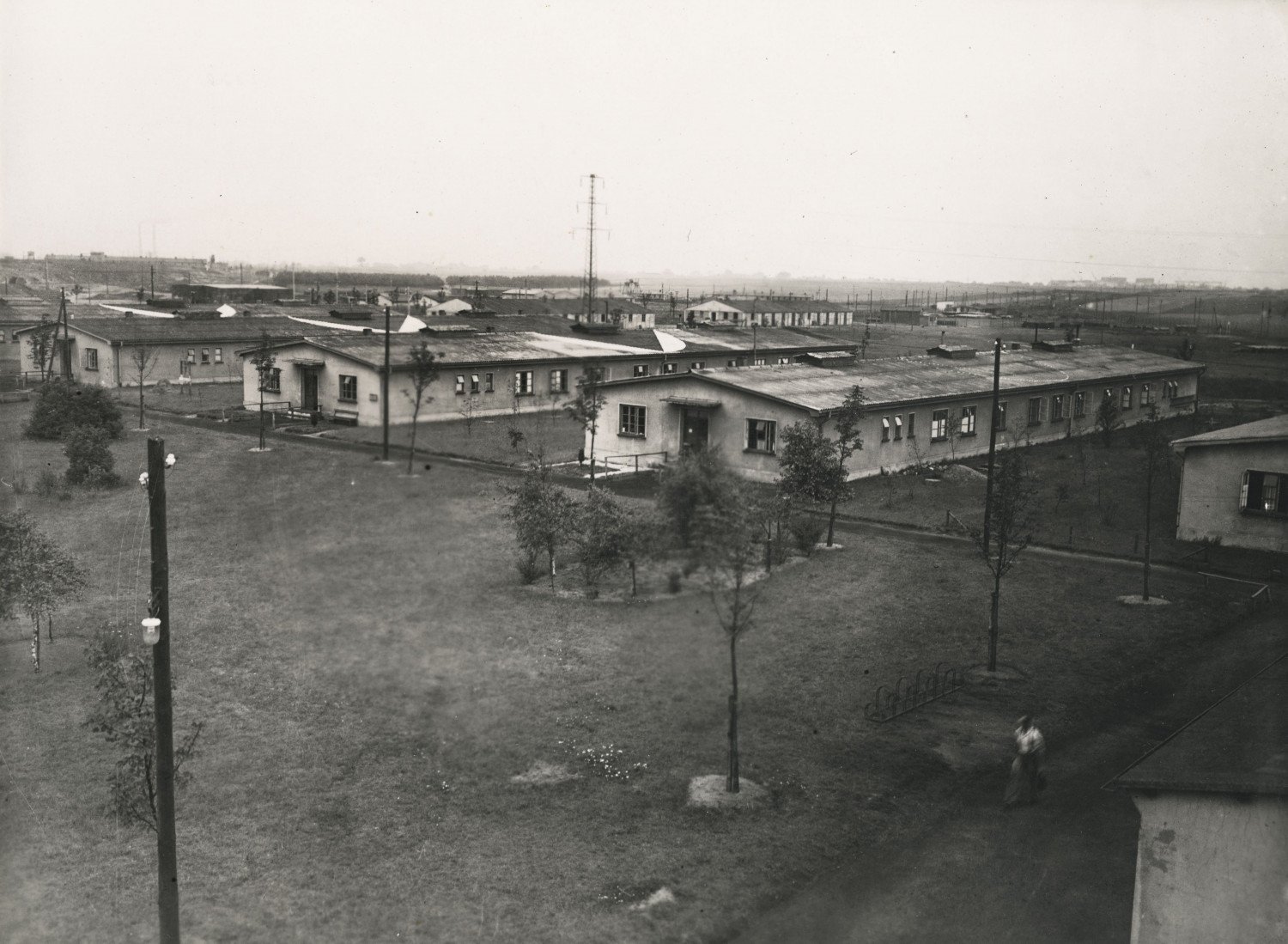 Gezeigt wird eine Luftaufnahme von mehreren Baracken. Im Vordergrund ist eine Wiese mit Bäumen zu sehen.  