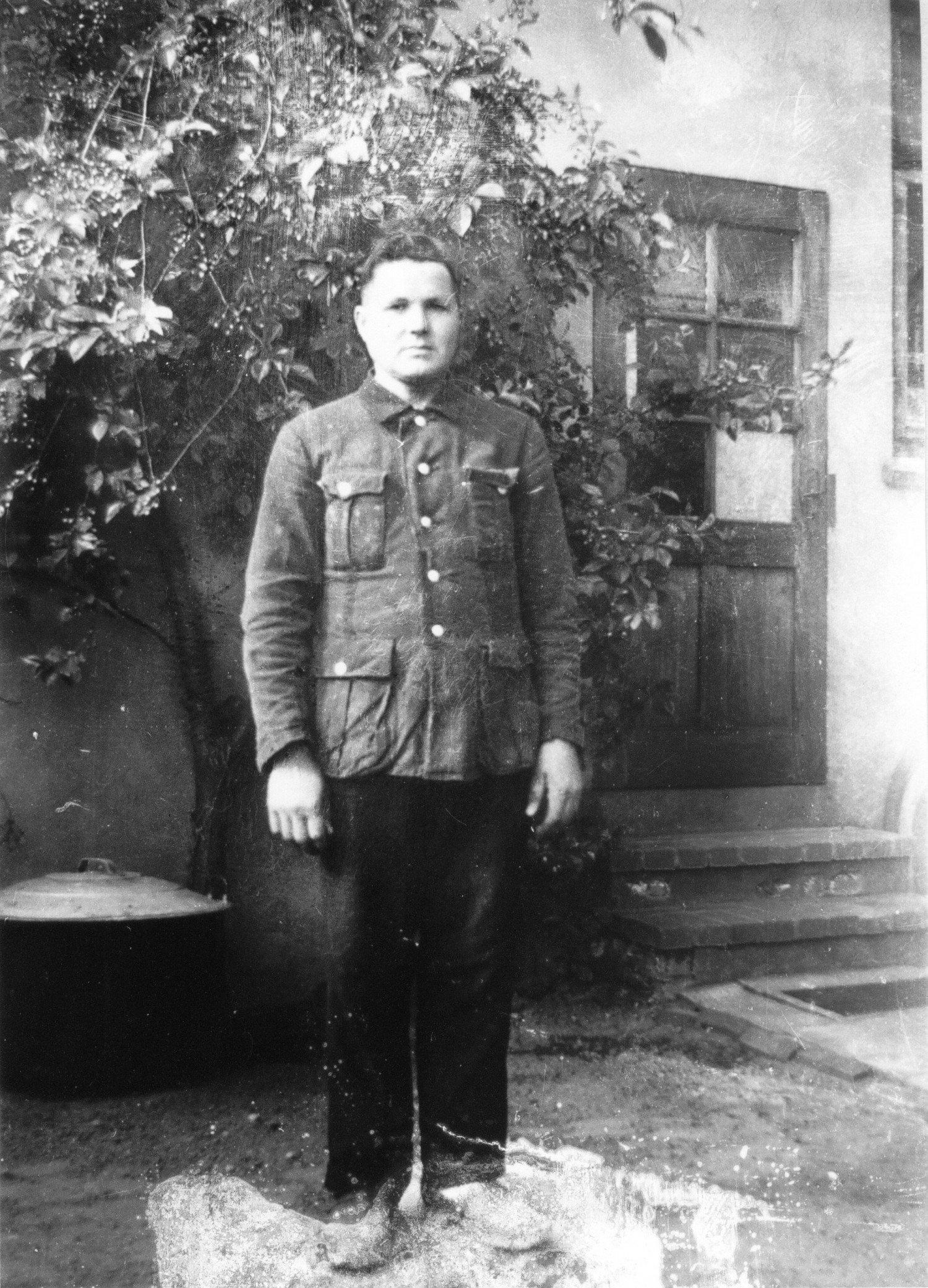 Abgebildet wird ein schwarz-weißes Bild, auf welchem ein Mann in Uniform steht. Er blickt in die Kamera. Im Hintergrund sieht man den Ausschnitt einer Haustür und einer Pflanze, die links neben der Tür an der Wand wächst. 