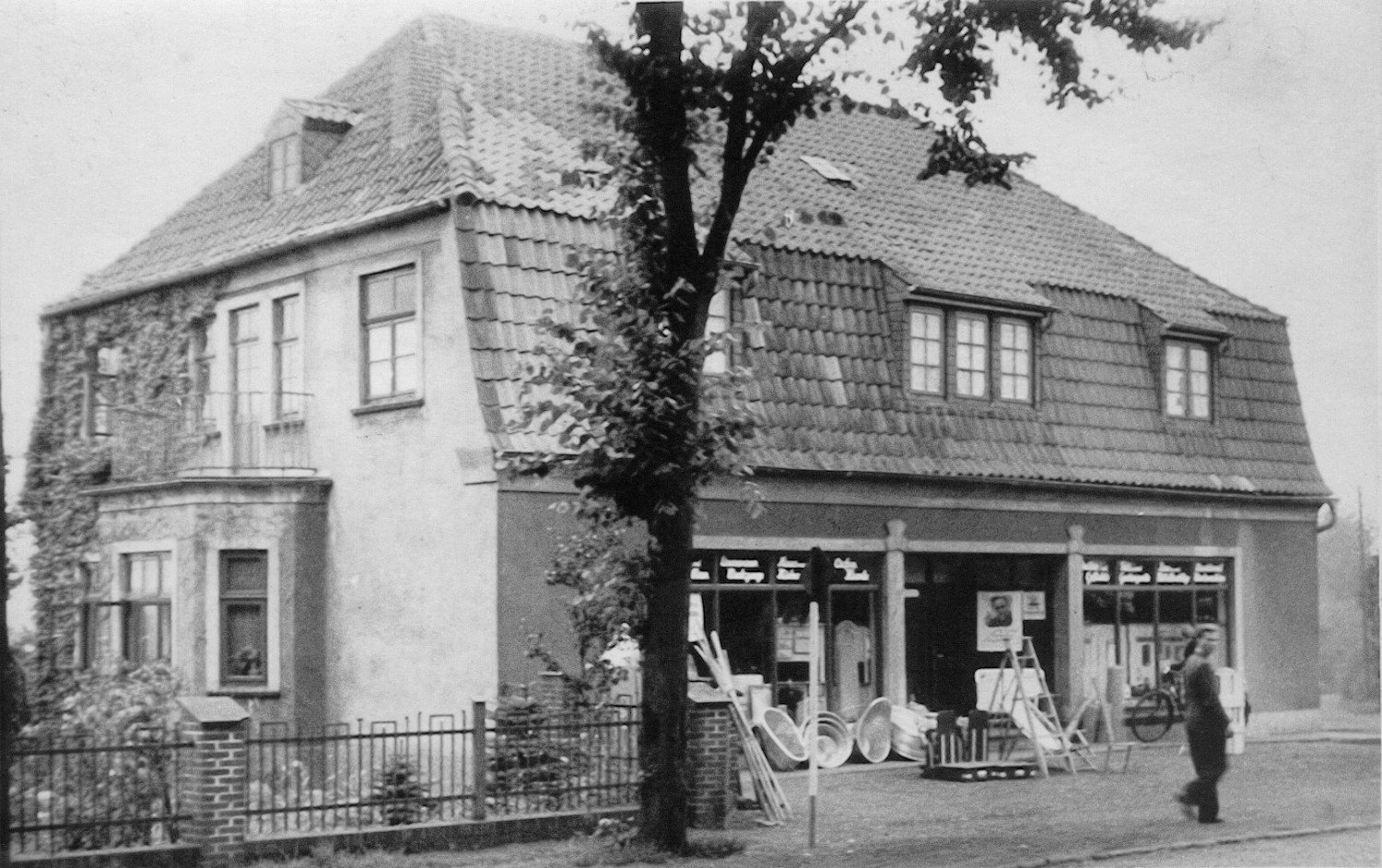 Gezeigt wird eine schwarz-weiß-Aufnahme von einem Haus, im Vordergrund ist eine laufende Frau sowie ein großer Baum zu sehen. 
