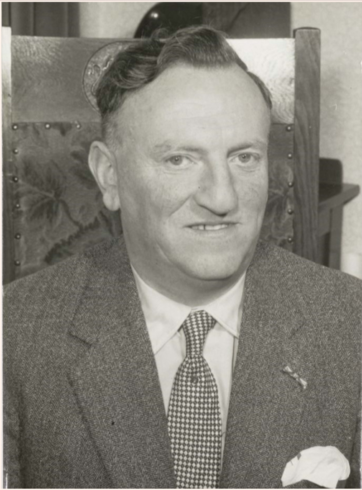 Auf dem schwarz-weißen Bild ist ein Mann in Anzug abgebildet. Er trägt ein Hemd und eine karierte Krawatte, in seinem Sakko steckt in der rechten Seite ein Tuch. Hinter ihm befindet sich ein mit Stoff überzogener Stuhl, auf dem er vermutlich sitzt. Die Lehne ist sehr hoch. Der Mann lächelt auf dem Foto etwas.   