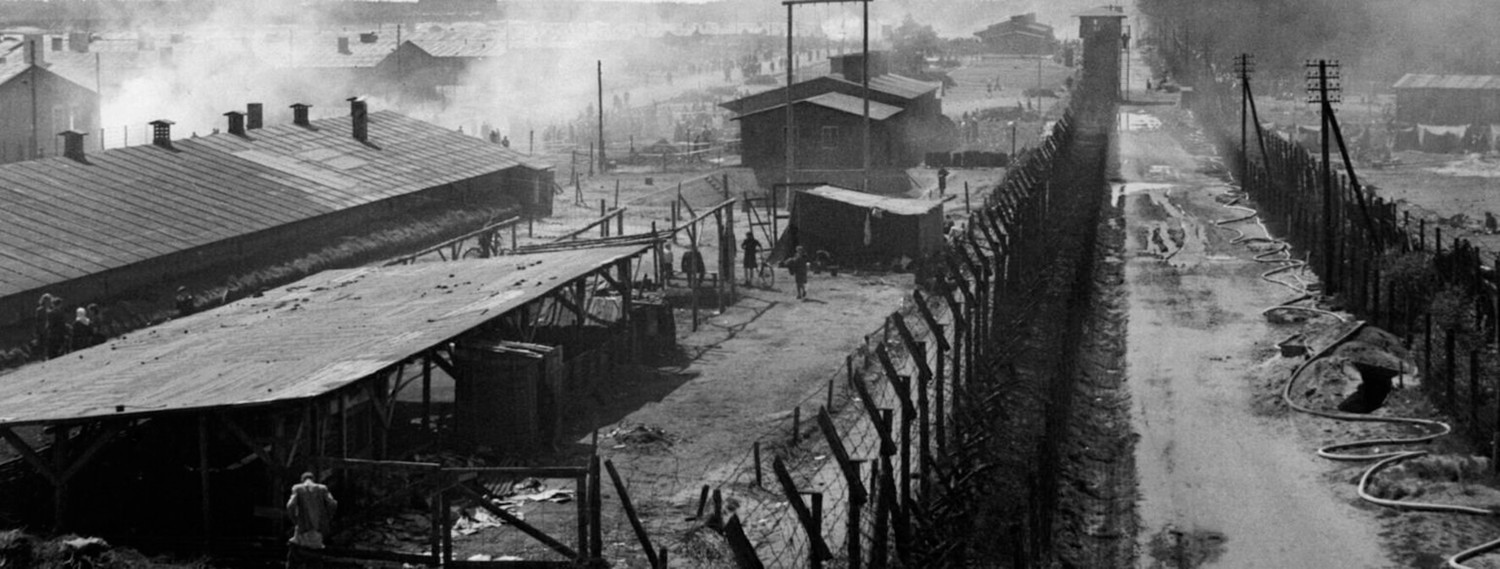 Auf dem schwarz-weißen Bild sind viele Holzunterkünfte mit Blechdächern und Kaminen eines Lagers abgebildet. Es handelt sich um das KZ Bergen-Belsen. Im Vordergrund sieht man einen Stacheldrahtzaun, der mit einem Wachturm versehen ist. Der Zaun bietet eine Art Durchgang oder Durchfahrtmöglichkeit. Auf der linken Seite des Zauns stehen Menschen, auf dem Boden liegt Kleidung und in der Luft befindet sich Rauch - es sieht aus, als würden Teile des Lagers brennen. In der vorderen, linken Ecke des Bildes sieht man einen Mann, der kein Oberteil anhat. Auf der rechten Seite des Bildes sieht man ebenfalls einen vermutlich brennenden Teil des Lagers mit hölzernen Unterkunftsgebäuden.  